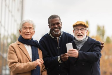 Akıllı telefonlu çok ırklı emekliler şehir sokağındaki kameraya bakıyor. 