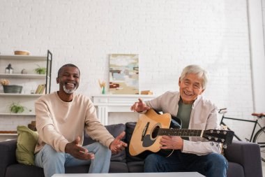 Akustik gitarlı neşeli çok ırklı emekliler evdeki kanepede kameraya bakıyor. 