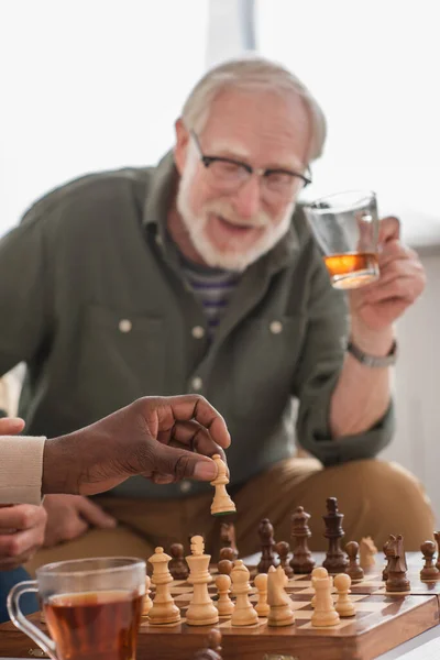 African american man holding chess figure near tea and blurred friends at home