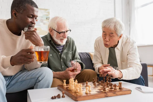 Interracial elderly friends holding tea while playing chess in living room