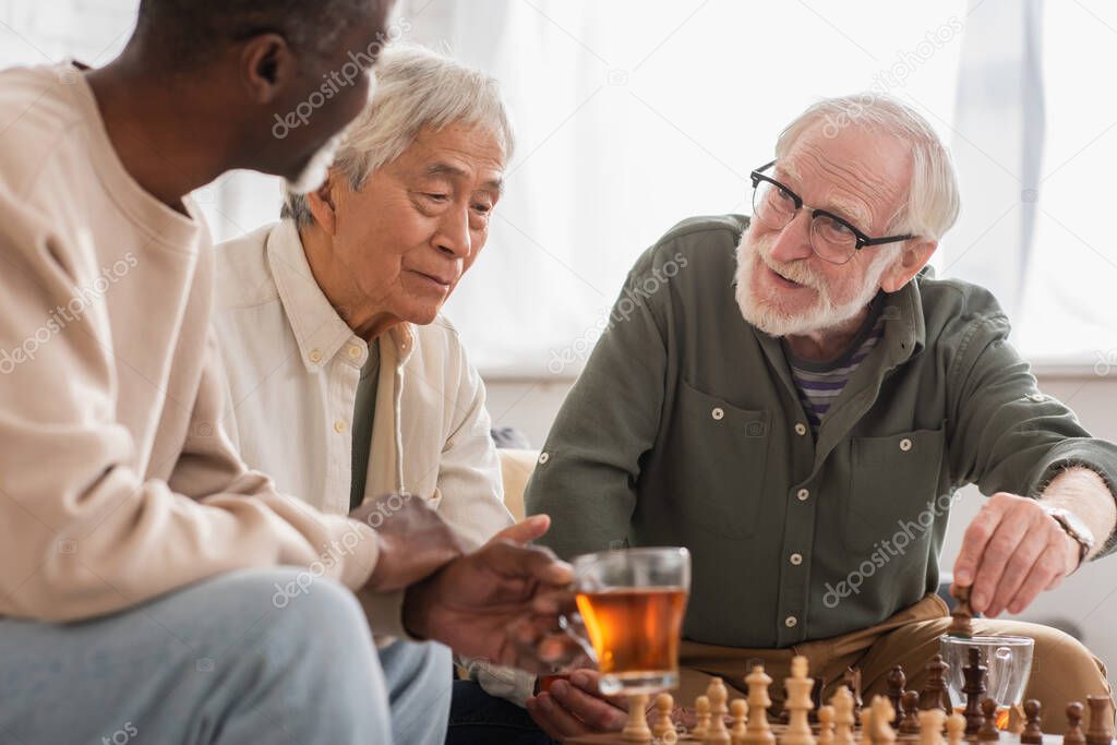 Smiling man playing chess with interracial friends near tea at home 