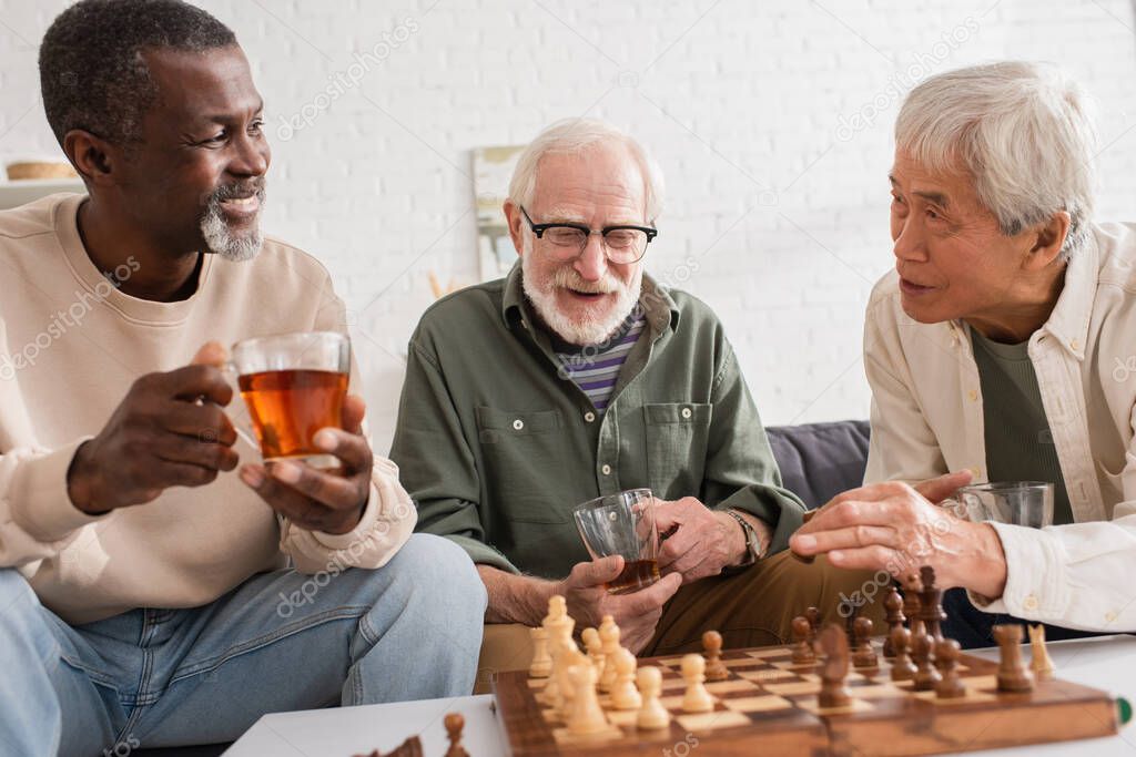Multicultural friends holding cups of tea near chessboard at home 