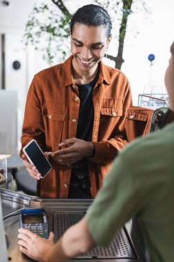 Gülümseyen Afro-Amerikan müşteri kafedeki bulanık baristanın yanında akıllı telefonuyla işaret ediyor. 