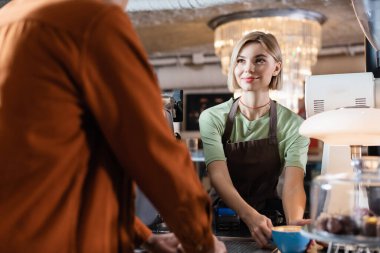Kafeteryadaki bulanık Afrikalı Amerikalı müşteriye yaklaşmak için önlüklü kahveyle gülümseyen barista 