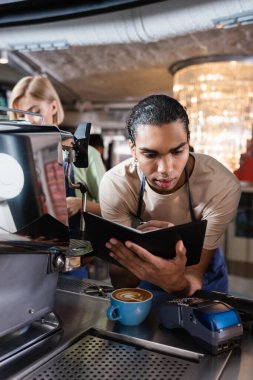 Kafedeki kahve ve ödeme terminalinin yakınındaki deftere yazı yazan Afrikalı Amerikalı barista. 