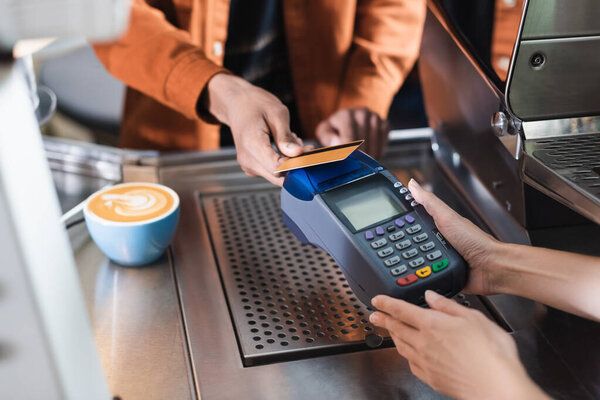 Cropped view of barista holding payment terminal near african american customer and coffee in cafe 