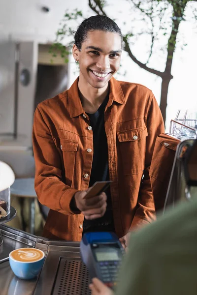 持信用卡的非洲裔美国人微笑着 带着付款终端和咖啡在模糊的咖啡店附近 — 图库照片