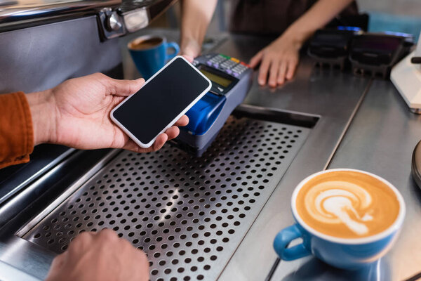 Cropped view of african american man holding cellphone near barista with payment terminal and coffee in cafe 