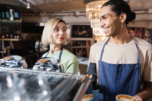 同僚とコーヒーマシンの近くにコーヒーを持っているアフリカ系アメリカ人のバリスタの笑顔カフェで — ストック写真