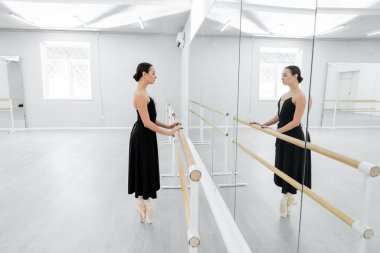 side view of ballet dancer in black dress standing on toes near barre in studio clipart