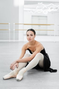 brunette ballerina looking at camera while sitting on floor in studio clipart