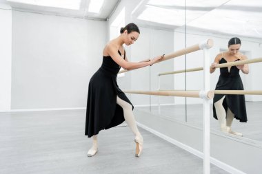 full length view of ballerina in black dress and pointe shoes training at barre near mirrors clipart