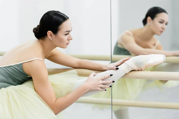brunette ballet dancer stretching leg at barre near mirrors in studio