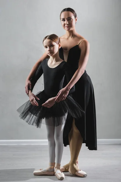 full length view of ballet teacher and girl in tutu looking at camera during rehearsal