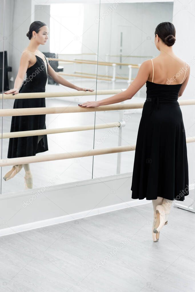 full length view of ballerina in black dress rehearsing near mirrors in studio