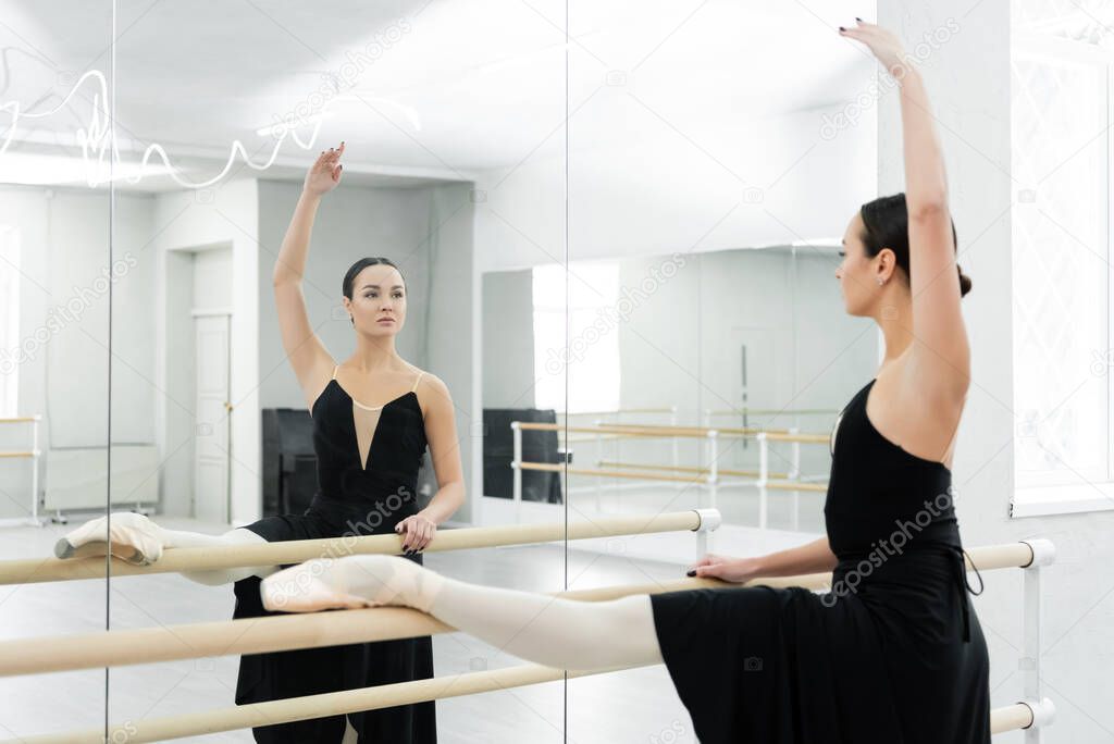 brunette ballerina looking in mirror while training at barre in dancing hall