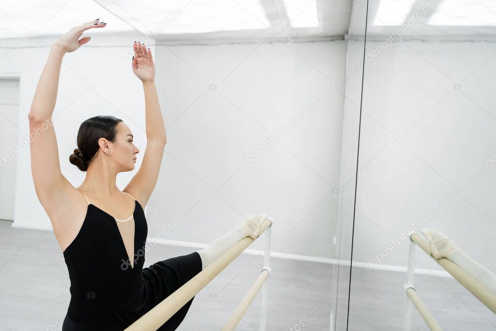 brunette woman with raised hands stretching at barre in ballet studio