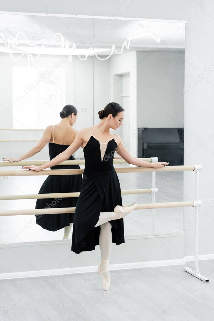 dancer in black dress exercising on barre near mirrors in ballet studio