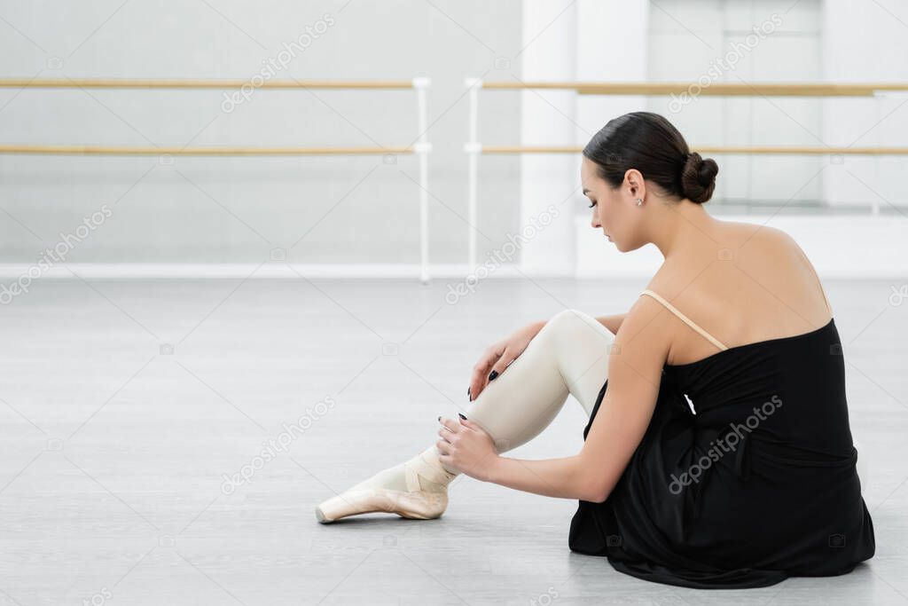 brunette ballerina adjusting pointe shoe while sitting on floor in studio