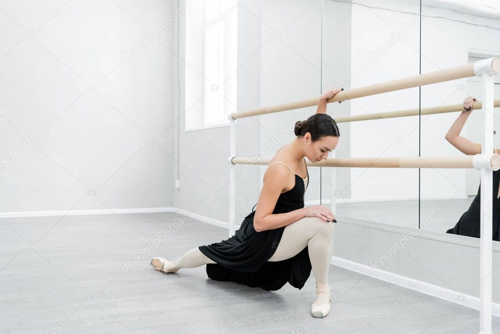 full length view of ballerina in black dress stretching at barre in dance studio