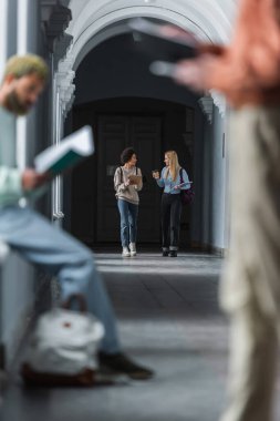 Kahveli ve dizüstü bilgisayarlı gülümseyen çok ırklı öğrenciler üniversite koridorunda yürüyorlar 