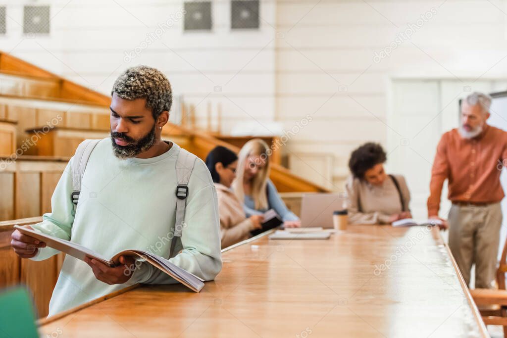 African american student holding notebook near blurred friends and teacher in university auditorium 