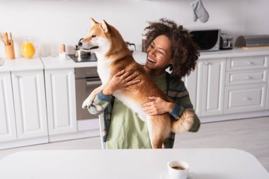 Heyecanlı Afrikalı Amerikalı kadın kahve fincanının yanında otururken Shiba Inu köpeği tutuyor.
