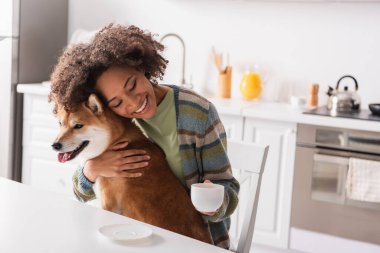 Mutfakta Shiba Inu köpeğini kucaklayan gözleri kapalı ve kahve fincanı olan mutlu Afrikalı Amerikalı kadın.