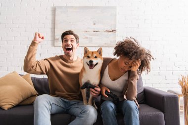 KYIV, UKRAINE - 22 Aralık 2021: Üzgün Afrikalı Amerikalı kız arkadaşı ve Shiba Inu 'nun yanında joystick tutan heyecanlı adam 