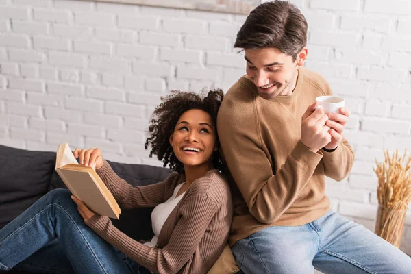 Hombre Sonriente Con Taza Mirando Novia Afroamericana Con Libro Sofá — Foto de Stock