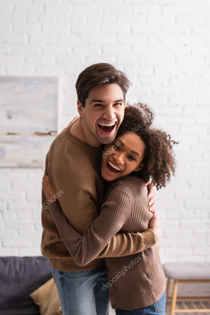 Positive multiethnic couple embracing and looking at camera at home 