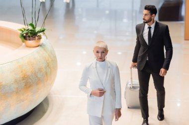 high angle view of senior businesswoman in white suit near bi-racial bodyguard with suitcase in hotel hall clipart
