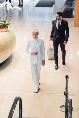 Otel lobisinde bavullu iki ırklı korumanın yanında yürüyen şık bir iş kadınının yüksek açılı görüntüsü.