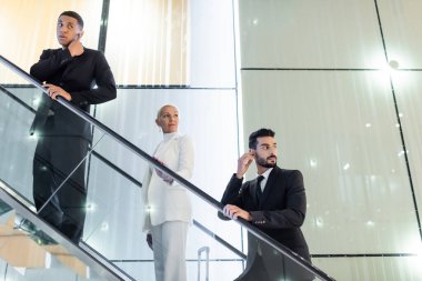 interracial bodyguards adjusting earpiece near wealthy senior businesswoman on hotel escalator clipart