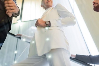 Beyaz elbiseli kıdemli iş kadınının, oteldeki yürüyen merdivende ırklar arası korumaların yanında alçak açılı görüntüsü.