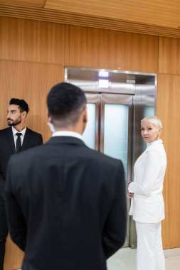 Çok ırklı güvenlik görevlilerinin yakınında, otelde asansör bekleyen şık bir üst düzey iş kadını.