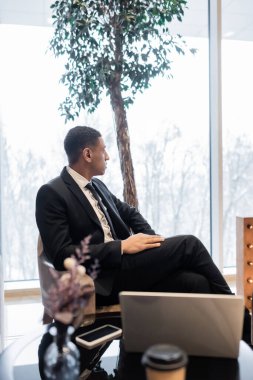Afro-Amerikan güvenlik görevlisi otel odasında bulanık dizüstü bilgisayarın ve cep telefonunun yanında oturuyor.