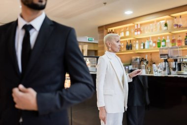 wealthy businesswoman near lobby bar and blurred mixed race bodyguard in hotel clipart