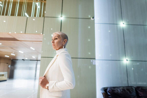 side view of blonde senior businesswoman in white blazer in lobby of modern hotel