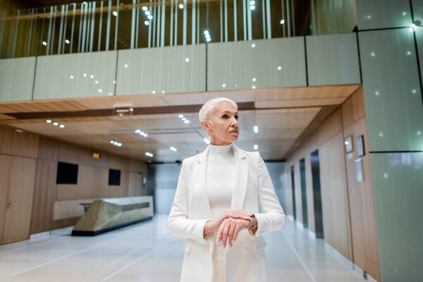 mature businesswoman in white trendy clothes looking away in lobby of modern hotel
