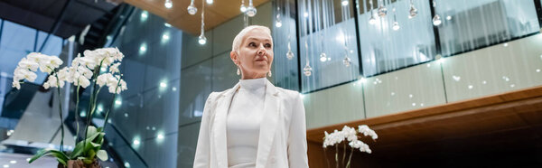 mature businesswoman in white blazer looking away near orchids in hotel lounge, banner