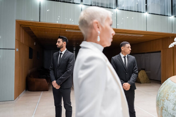 interracial bodyguards in suits protecting senior business lady in hotel on blurred foreground