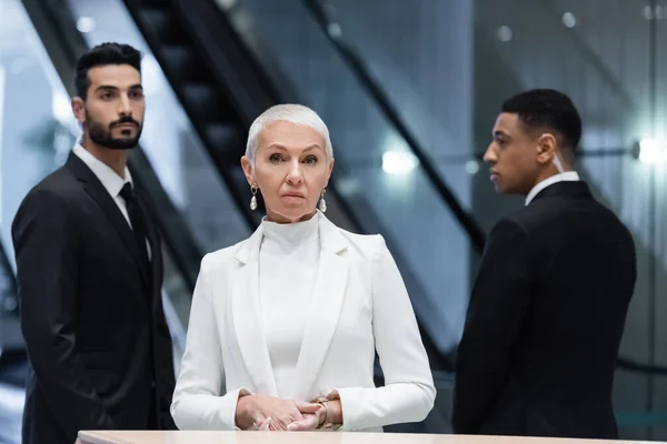 mature and stylish business lady standing at hotel reception near interracial bodyguards