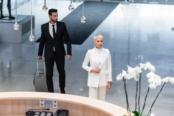 high angle view of stylish senior business lady and private bi-racial bodyguard with travel bag near reception of luxury hotel