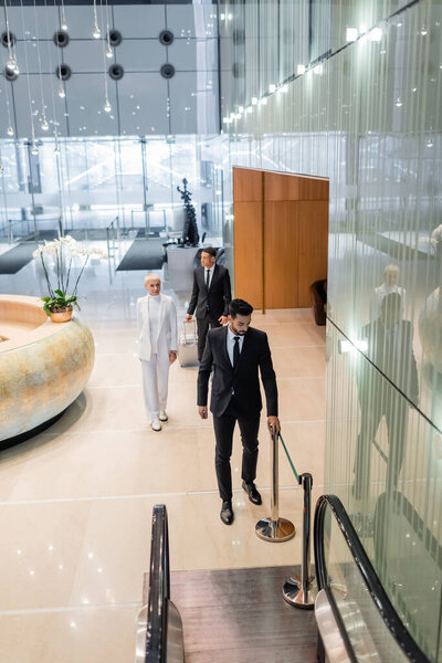 высокий угол зрения пожилой предпринимательницы с межрасовыми телохранителями, идущей к эскалатору отеля 