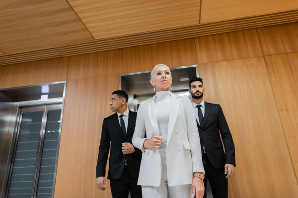 low angle view of successful mature business lady near multiethnic bodyguards in hotel hall