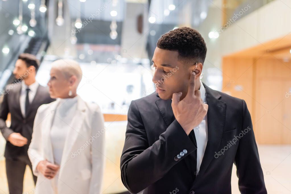 african american bodyguard adjusting earpiece near mature businesswoman with bi-racial guard on blurred background