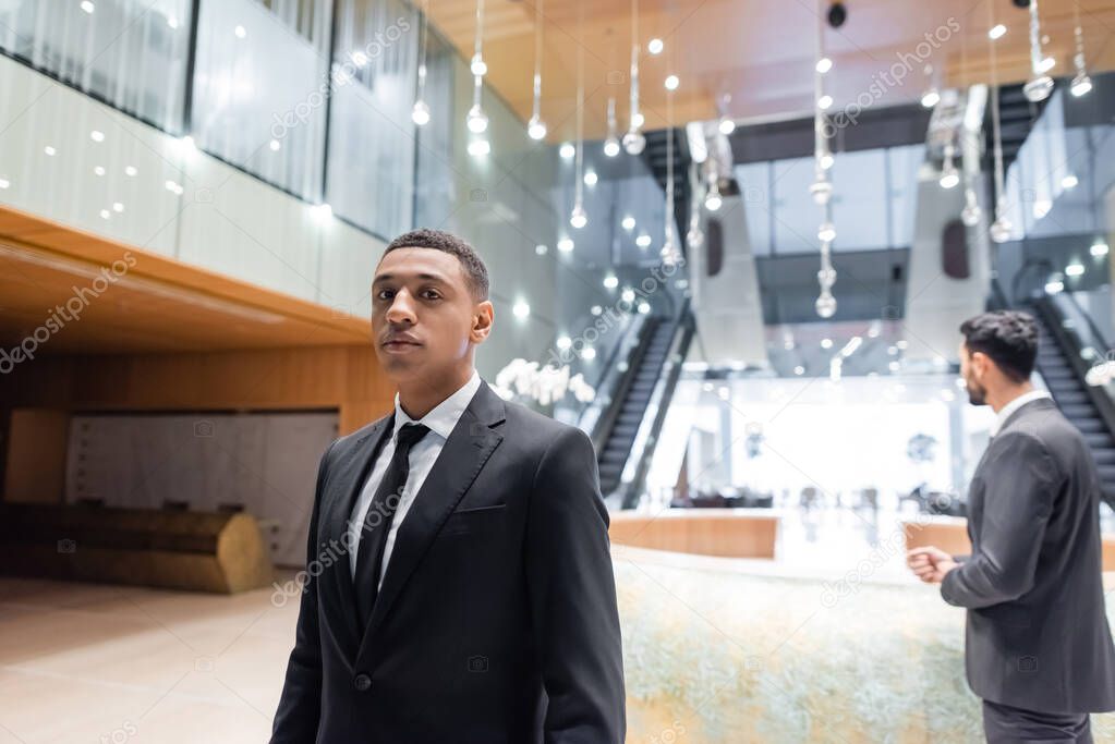 african american security man looking at camera near blurred bi-racial colleague at hotel reception