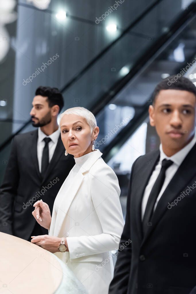 stylish senior businesswoman looking away near private interracial bodyguards in hotel lobby