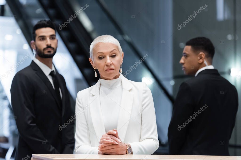 stylish senior business lady standing at hotel reception near blurred interracial bodyguards